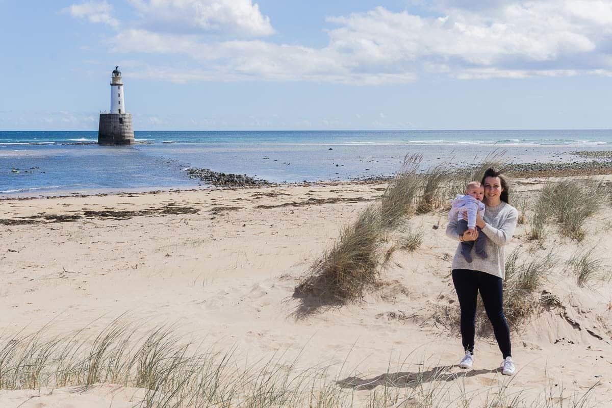 Holding baby on the beach with lighthouse in the background.